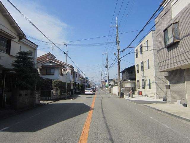 町の西端を走る主要道路を南方向から見た景色。沿道には一戸建てや店舗などが見られます。